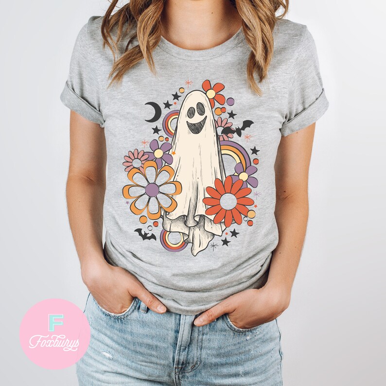 Peut inclure: Un t-shirt gris avec un fant&ocirc;me blanc souriant entour&eacute; de fleurs color&eacute;es, d'&eacute;toiles et de chauves-souris. Le design a une ambiance r&eacute;tro des ann&eacute;es 70.
