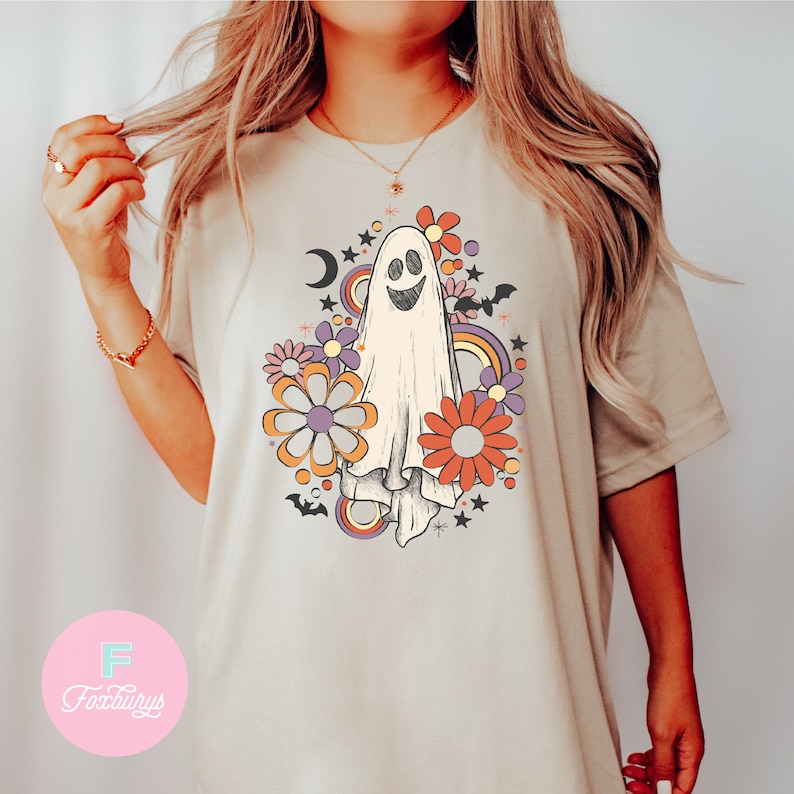 Peut inclure: Un t-shirt beige avec un fant&ocirc;me blanc souriant entour&eacute; de fleurs color&eacute;es et d'accents arc-en-ciel. Le fant&ocirc;me est entour&eacute; d'&eacute;toiles et de chauves-souris.