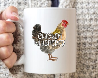 May include: White ceramic mug featuring a watercolor illustration of a chicken with the text "Chicken Whisperer". The chicken has brown, black, and yellow feathers, a red comb, and a yellow beak. The mug has a white handle.