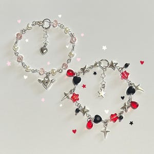 May include: Two silver charm bracelets with different themes. One bracelet features pink and white beads with a silver heart charm and a silver angel wing charm. The other bracelet features red and black beads with a silver star charm, a silver heart charm, and a silver cross charm.