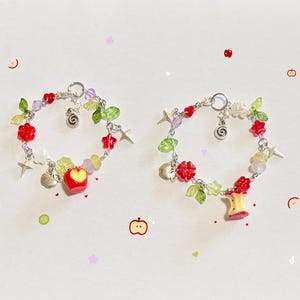 May include: Two circular charm bracelets featuring silver-tone chains adorned with red apple-shaped beads, green leaf-shaped beads, small star charms, and translucent purple and yellow beads. One bracelet includes a whole apple charm, while the other features an apple core charm and a small silver spiral charm.