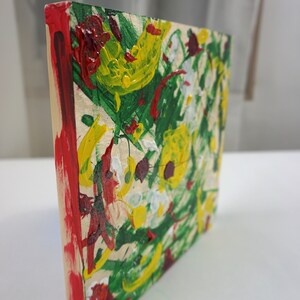 May include: A small, square wooden canvas with a colorful abstract painting. The painting features a mix of green, yellow, red, and white paint, creating a vibrant and energetic design.