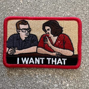 May include: Embroidered patch featuring a couple at a table, with the text "I WANT THAT" in white. The patch has a red border, a tan background, and depicts a man in glasses and a woman in a red top.
