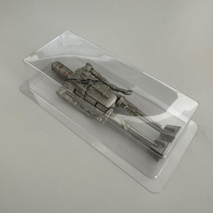 May include: A silver-colored droid action figure with a slender, elongated frame and a rounded head, encased in a clear, rectangular plastic blister pack. The figure features articulated limbs and detailed mechanical components on its torso.