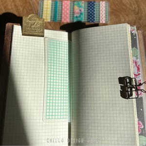 Peut inclure: Carnet ouvert avec du papier quadrill&eacute;, un signet quadrill&eacute; vert clair et un trombone en laiton. Une bande d&eacute;corative de washi tape avec divers motifs est au-dessus du carnet. Le carnet a un trombone en laiton qui dit "Traveler's Notebook". Le texte "CHELLO DESIGN SHOP" est en bas.