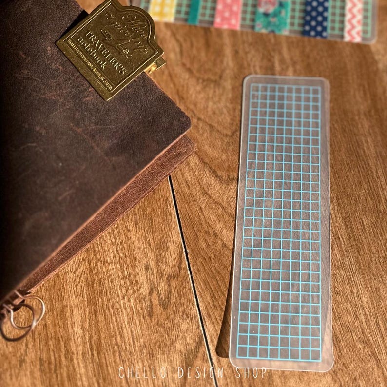 Peut inclure: Un marque-page rectangulaire transparent avec un motif de grille bleu. Un carnet en cuir marron avec un clip en m&eacute;tal dor&eacute; est visible. Le marque-page est sur une surface en bois.