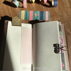 Peut inclure: Un carnet ouvert avec du papier quadrill&eacute;, une r&egrave;gle transparente et une pince m&eacute;tallique. Des rouleaux de washi tape d&eacute;coratifs et une bande de ruban adh&eacute;sif &agrave; motifs sont dispos&eacute;s au-dessus du carnet. Un signet en laiton est fix&eacute; en haut du carnet. Le texte "CHELLO DESIGN SHOP" est en bas.