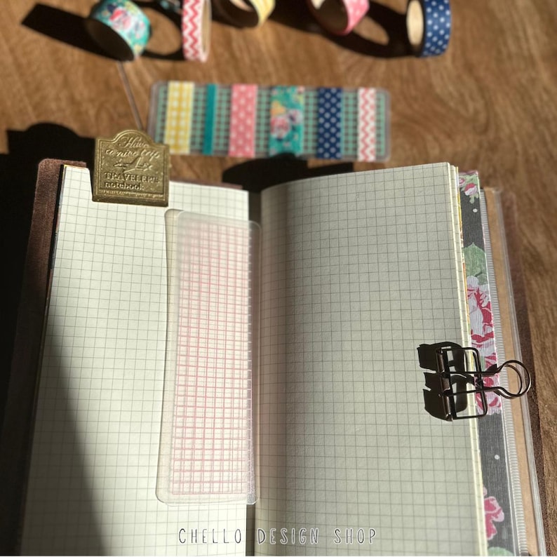Peut inclure: Un carnet ouvert avec du papier quadrill&eacute;, une r&egrave;gle transparente et une pince m&eacute;tallique. Des rouleaux de washi tape d&eacute;coratifs et une bande de ruban adh&eacute;sif &agrave; motifs sont dispos&eacute;s au-dessus du carnet. Un signet en laiton est fix&eacute; en haut du carnet. Le texte "CHELLO DESIGN SHOP" est en bas.
