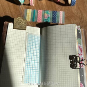 Peut inclure: Carnet ouvert avec du papier quadrill&eacute;, un signet transparent &agrave; quadrillage bleu et une pince m&eacute;tallique dor&eacute;e. Des rouleaux de washi tape d&eacute;coratifs sont en arri&egrave;re-plan. Le texte "HELLO DESIGN SHOP" est visible en bas de la page.
