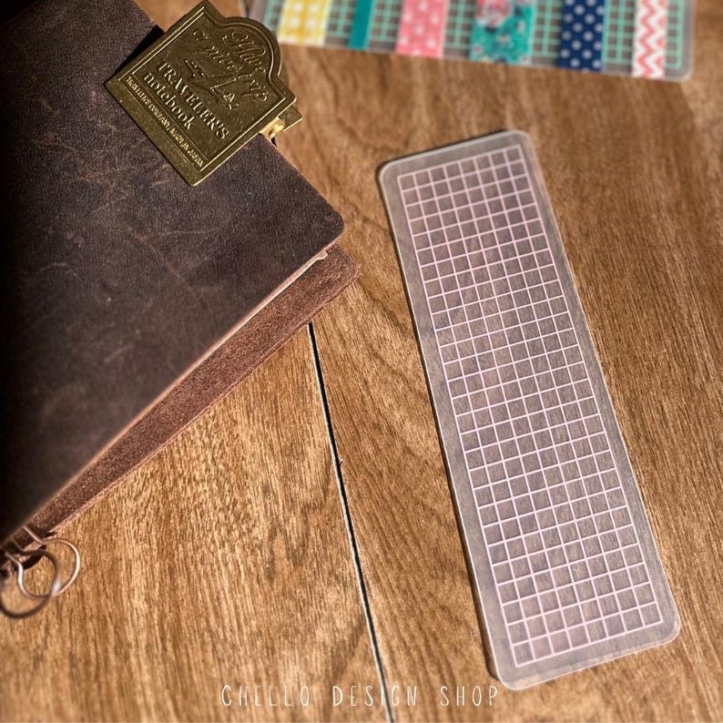 Peut inclure: Un marque-page rectangulaire transparent avec un motif quadrill&eacute; rose clair. Il est pos&eacute; &agrave; c&ocirc;t&eacute; d'un carnet en cuir marron avec un clip m&eacute;tallique dor&eacute;. Le clip du carnet porte l'inscription "Traveler's Notebook".
