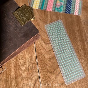 Peut inclure: Marque-page en plastique transparent avec un motif quadrill&eacute; bleu clair. Le marque-page est rectangulaire et bord&eacute;. Un carnet en cuir marron avec un clip en m&eacute;tal dor&eacute; est en arri&egrave;re-plan. Le clip indique "Traveler's Notebook".