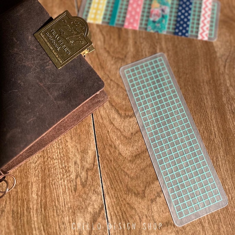 Peut inclure: Marque-page en plastique transparent avec un motif quadrill&eacute; bleu clair. Le marque-page est rectangulaire et bord&eacute;. Un carnet en cuir marron avec un clip en m&eacute;tal dor&eacute; est en arri&egrave;re-plan. Le clip indique "Traveler's Notebook".