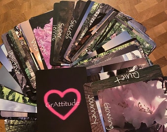 May include: A set of affirmation cards with various nature scenes and positive words like "Success", "Super", "Upbeat", "Journey", "Glowing", "Gratitude", and "Out",  arranged in a fan shape on a wooden surface.