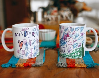May include: Two white ceramic mugs with colorful drawings on them. The mug on the left says "Amber" and the mug on the right has a drawing of a person with a smiley face. Both mugs are sitting on colorful woven placemats.