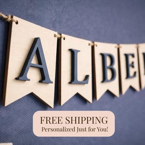 May include: A wooden banner spelling "ALBERT" with navy blue letters, strung on twine. The banner is made of light-colored wood with a decorative edge. A string of wooden beads hangs on the left side. The background is a textured blue surface.
