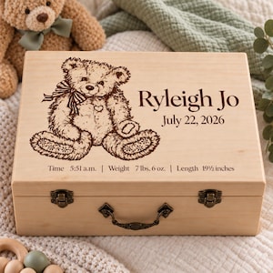May include: A rectangular light-colored wooden keepsake box featuring a laser-engraved teddy bear, the name Ryleigh Jo, the date July 22, 2026, and birth details: Time 5:51 a.m., Weight 7 lbs. 6 oz., and Length 19 1/2 inches, with metal latches.
