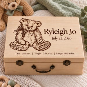 May include: A rectangular light-colored wooden keepsake box with a hinged lid, featuring a laser-engraved teddy bear, the name Ryleigh Jo, the date July 22, 2026, and birth details: Time 5:51 a.m., Weight 7 lbs. 6 oz., and Length 19 1/2 inches.