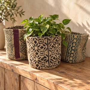 May include: Three cylindrical woven baskets made of natural jute fibre, each featuring distinct dark blue and black geometric patterns. The centre basket holds a vibrant green pothos plant with heart-shaped leaves, while the others remain empty.