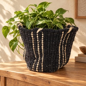 May include: A black woven fabric basket with vertical cream-coloured stripes containing a green pothos plant with variegated heart-shaped leaves. The basket features a textured, hand-woven construction with a wide opening and a soft, flexible structure.