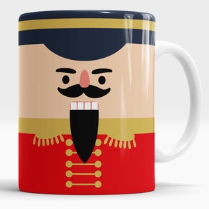 May include: White ceramic mug with a colourful illustration of a nutcracker soldier's face. The design includes a blue hat with gold trim, a black moustache, and a red jacket with gold buttons.