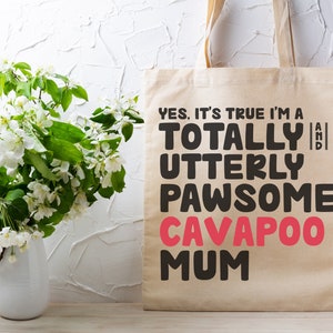 May include: A beige canvas tote bag with the text "YES, IT'S TRUE I'M A TOTALLY AND UTTERLY PAWSOME CAVAPOO MUM" in black and pink lettering. A vase of white flowers is to the left.