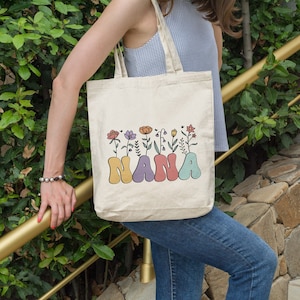 May include: A white canvas tote bag featuring the word "NANA" in colourful, stylised letters. The letters are adorned with small flowers in a variety of colours.