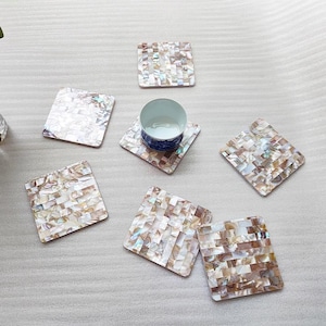 May include: A set of square coasters with a mosaic design made from iridescent shell pieces. The coasters are in shades of white, beige, and brown, with a glossy finish. A small blue and white patterned cup sits on one of the coasters.
