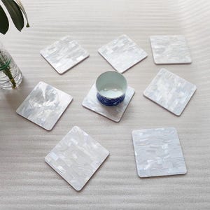 May include: A collection of square, white coasters with a mother-of-pearl effect. One coaster holds a small, blue and white patterned cup. The coasters are arranged on a light gray surface, with a vase of greenery in the background.
