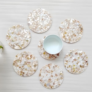 May include: A set of seven round coasters with a mosaic design made of iridescent, natural-colored shell pieces. One coaster holds a light blue ceramic cup. The coasters are arranged on a light-colored surface.