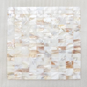 Może przedstawiać: Kwadratowa płytka mozaikowa złożona z małych, prostokątnych kawałków. Płytki mają perłową, opalizującą jakość z odcieniami bieli, kremu i subtelnymi nutami brzoskwini i brązu. Mozaika ułożona jest w wzór kratki, tworząc teksturowaną powierzchnię.