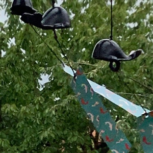 Puede incluir: Campanas de viento con forma de ballena de metal negro colgadas de cuerdas negras. Las campanas tienen una cola curvada y un anillo para colgar. Banderines de tela coloridos con diseños de peces son visibles en primer plano, con follaje verde en el fondo.