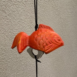 Puede incluir: Un adorno en forma de pez naranja brillante, suspendido por un cordón negro. El pez tiene aletas y escamas detalladas, y un accesorio de metal plateado. El fondo es una pared gris claro texturizada.