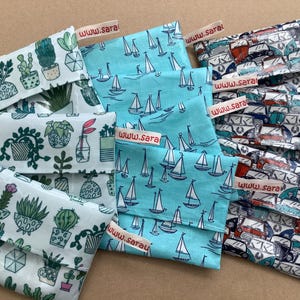 Könnte beinhalten: Mehrere Stoffbeutel mit verschiedenen Mustern werden gezeigt. Einer hat ein Kaktus- und Topfpflanzen-Design, ein anderer ein Segelbootmuster und ein dritter einen Vintage-Van-Druck. Jeder Beutel hat ein Etikett mit der Aufschrift "www.sara".