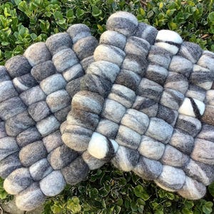 Può includere: Due sottopentola in feltro grigio e bianco con un design intrecciato. I sottopentola sono realizzati con pezzi di feltro cilindrici arrotondati in tonalità di grigio, bianco e marrone. I sottopentola sono posizionati su un letto di fogliame verde.