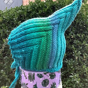 Pode incluir: Um chapéu de malha azul-esverdeado, verde e azul com um topo pontudo e uma longa gravata. O chapéu está sobre uma superfície com padrão roxo e branco.