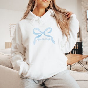 May include: White hooded sweatshirt with a blue watercolor bow and the text "Alpha Chi Omega" on the front.