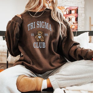 May include: Brown sweatshirt with a blue and white graphic of a teddy bear holding a heart. The graphic says "Tri Sigma 18 98 Club".