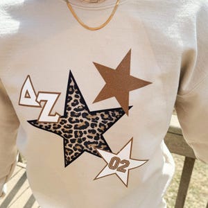 May include: A beige sweatshirt with a leopard print star, a brown star, and a white star with the number "02" on it. The letters "AZ" are on the leopard print star.