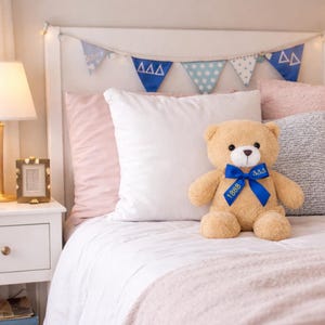 May include: A tan teddy bear with a blue ribbon and the text "1888" and "ΔΔΔ" is centered on a bed with white bedding and pink pillows. A banner with blue triangles and polka dots hangs above the bed.