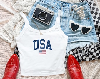 May include: A white tank top featuring the text USA in navy blue block letters above a small American flag graphic. The tank top is surrounded by light blue denim shorts, red cowboy boots, a black instant camera, white heart-shaped sunglasses, and a white hair clip.