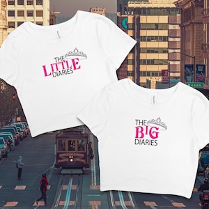May include: Two white crop tops with the text "The Little Diaries" and "The Big Diaries" in pink letters with a silver crown above each phrase.