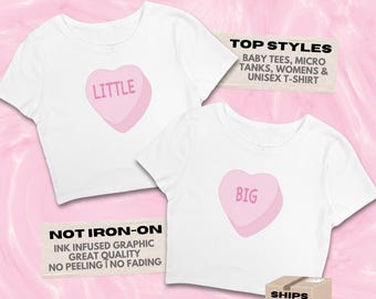 May include: Two white short-sleeved shirts featuring pink heart-shaped candy graphics. One shirt displays the word LITTLE, and the other displays the word BIG. Text on the image includes MY LITTLE SWEETHEART, TOP STYLES, NOT IRON-ON, and STIXBYLEX.