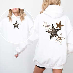 May include: White hoodie with a brown star with a leopard print design and the letters "AEA" in white on the back. A smaller brown star with the number "93" is on the bottom right. A smaller star with the letters "AEA" in white is on the front.