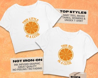 May include: Two white short-sleeved shirts featuring orange slice graphics. The left shirt displays the text THIS LITTLE IS A CUTIE, while the right shirt displays THIS BIG IS A CUTIE. The image includes text MY LITTLE IS A CUTIE and product details.