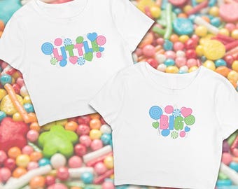 May include: Two white crop tops with short sleeves. The top on the left has the word "LITTLE" in colorful letters surrounded by candy-themed graphics. The top on the right has the word "BIG" in colorful letters surrounded by candy-themed graphics.