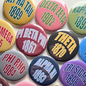 May include: A collection of round buttons with sorority and fraternity Greek letters and founding years. The buttons feature various colors, including yellow, pink, green, red, black, blue, and purple, with white polka dots. Each button displays Greek letters and a year.