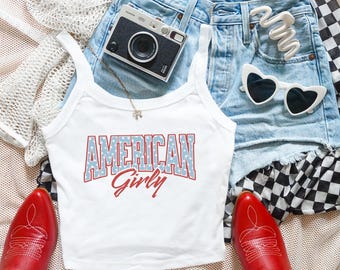 May include: A white tank top with the text AMERICAN GIRLY in blue with white stars and red outlines. The outfit includes light blue denim shorts, red cowboy boots, white heart-shaped sunglasses, a white hair clip, and a vintage film camera.