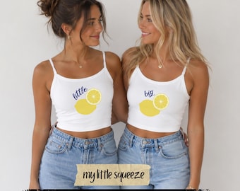 May include: Two white spaghetti strap crop tops featuring a yellow lemon graphic and the text "little" on one and "big" on the other. The tops are paired with light blue denim jeans. The design includes a yellow lemon slice and a whole lemon.
