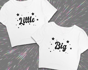 May include: Two white short-sleeved crop tops featuring black graphic text and star accents. One shirt displays the word Little in a cursive font, while the other displays the word Big in the same style, both surrounded by various sized black stars.