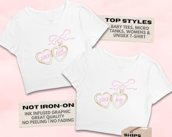 Könnte beinhalten: Zwei weiße Crop-Tops mit rosa Schleife und Herzanhänger-Design. Der Anhänger hat die Worte "xoxo little" und "xoxo big" in Gold. Der Text "CLOSE TO MY HEART" steht oben.